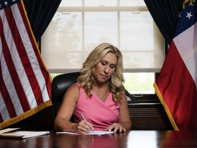 A woman in a pink sleeveless dress seated at a desk, writing on paper in a formal setting—symbolizing reflection and decision-making amid discussions surrounding Marjorie Taylor Greene resignation.